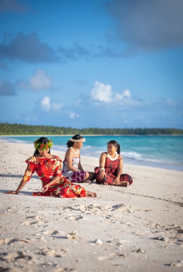 Three girls in bright dresses and flower crowns sitting on a white-sand beach by the lagoon